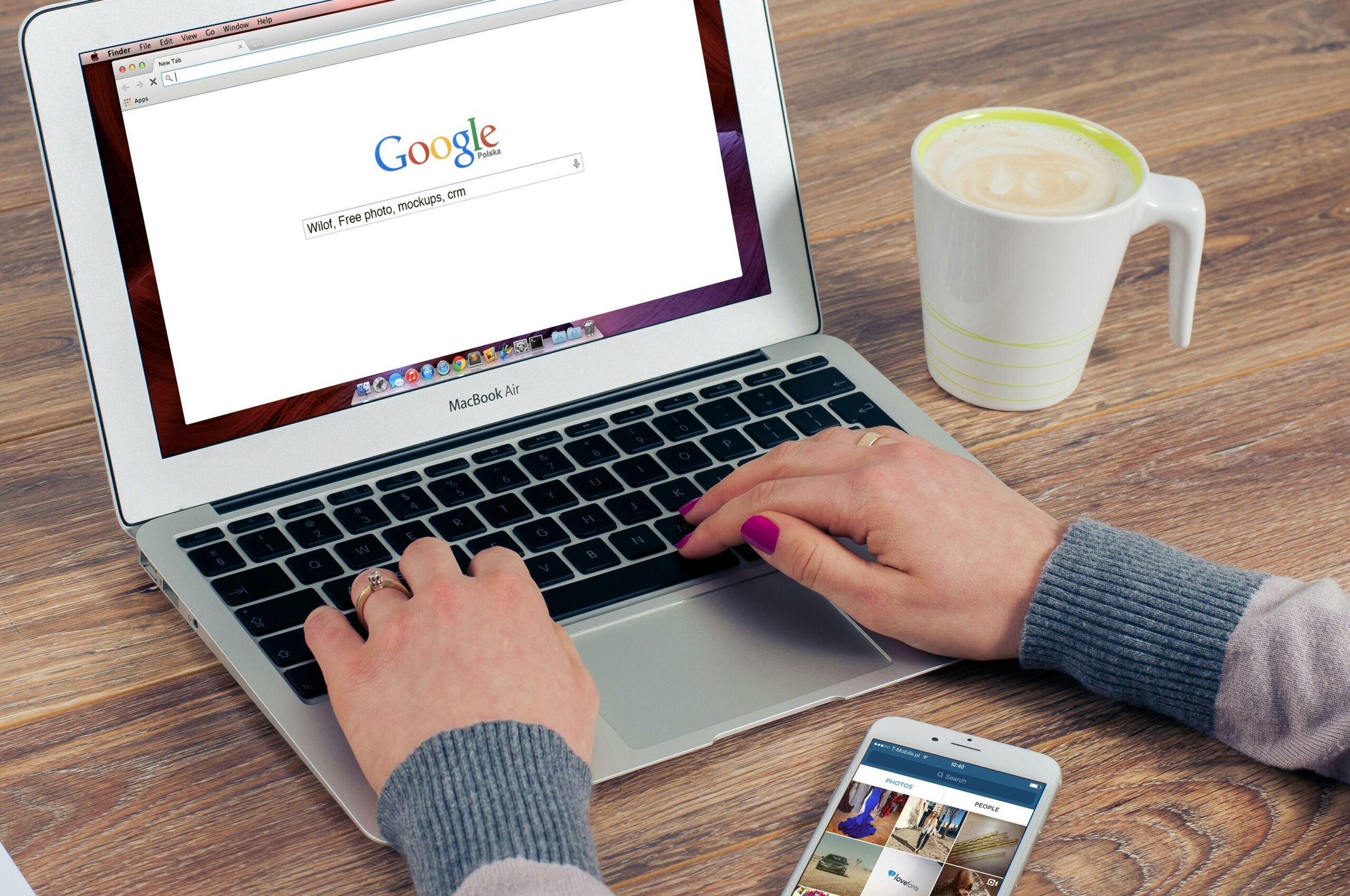 Hands typing on MacBook Air with Google search open, coffee nearby.